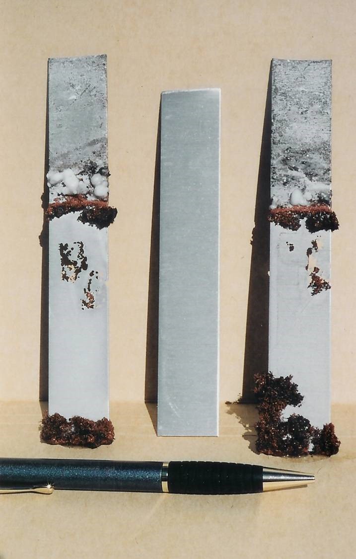 FIGURE 4 Two test strips of aluminum after exposure to a copper sulfate solution for several weeks. Note dark brown deposits of metallic copper on left and right strips. These strips are severely thinned and perforated. Center strip not exposed for comparison.